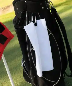 A Microfibre Golf Towel - TC19 hangs from a black golf bag near a red and black chequered flag on a green golf course. | Work & Wear DIrect