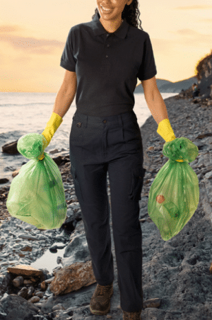 A person in dark clothing and yellow gloves walks along a rocky beach, smiling whilst holding two green rubbish bags filled with litter. The sun is setting over the sea in the background. | Work & Wear DIrect