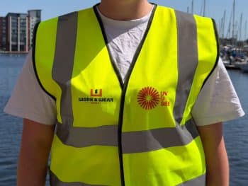 Person wearing a yellow high-visibility vest with WORK & WEAR and UV YOU SEE logos, standing by a marina with boats and buildings in the background. | Work & Wear DIrect