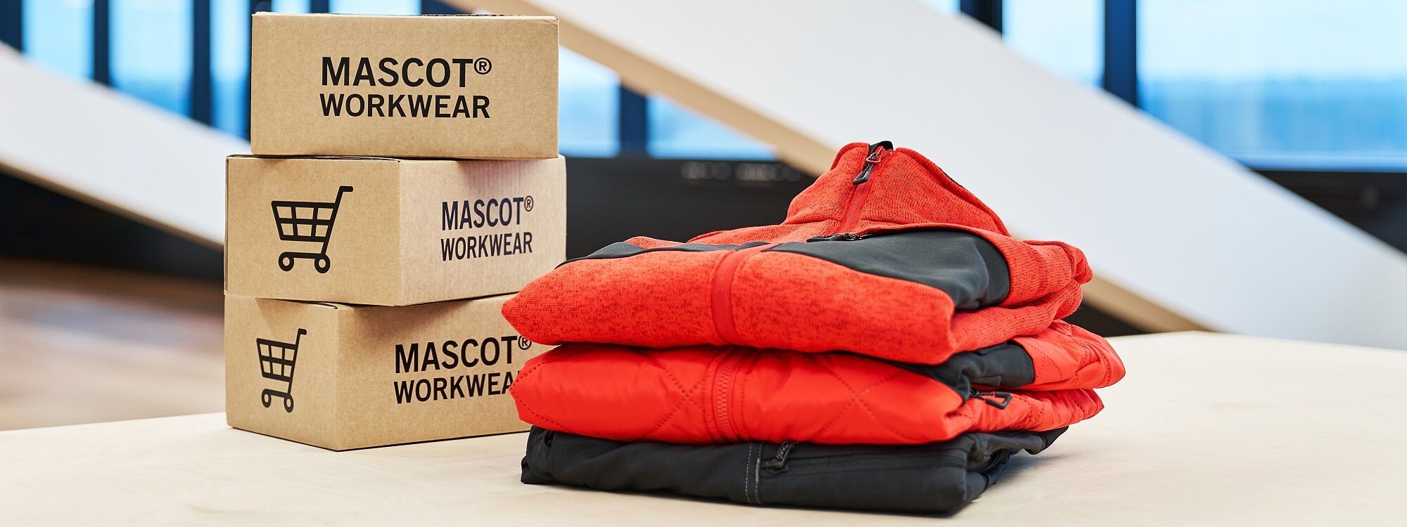 Three cardboard boxes labelled Mascot Workwear are stacked beside a neatly folded pile of red and black jackets on a light-coloured table, with large windows blurred in the background. | Work & Wear DIrect