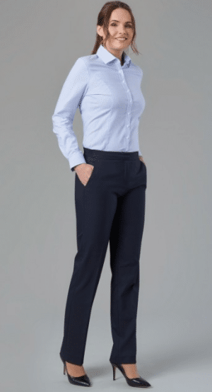 A woman stands smiling, wearing a light blue button-up shirt, dark dress trousers, and black heels with her hands in her pockets against a plain grey background. | Work & Wear DIrect